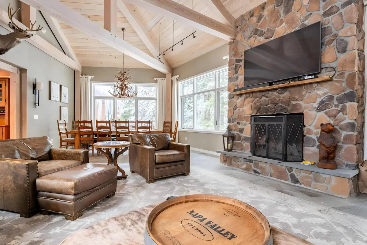 A warm, inviting interior scene featuring a striking stone fireplace as a focal point, with leather armchairs, a wooden dining table, and a blend of dining and living spaces. Large windows allow natural light to pour in, and the furnishings are arranged to foster conversation and togetherness.