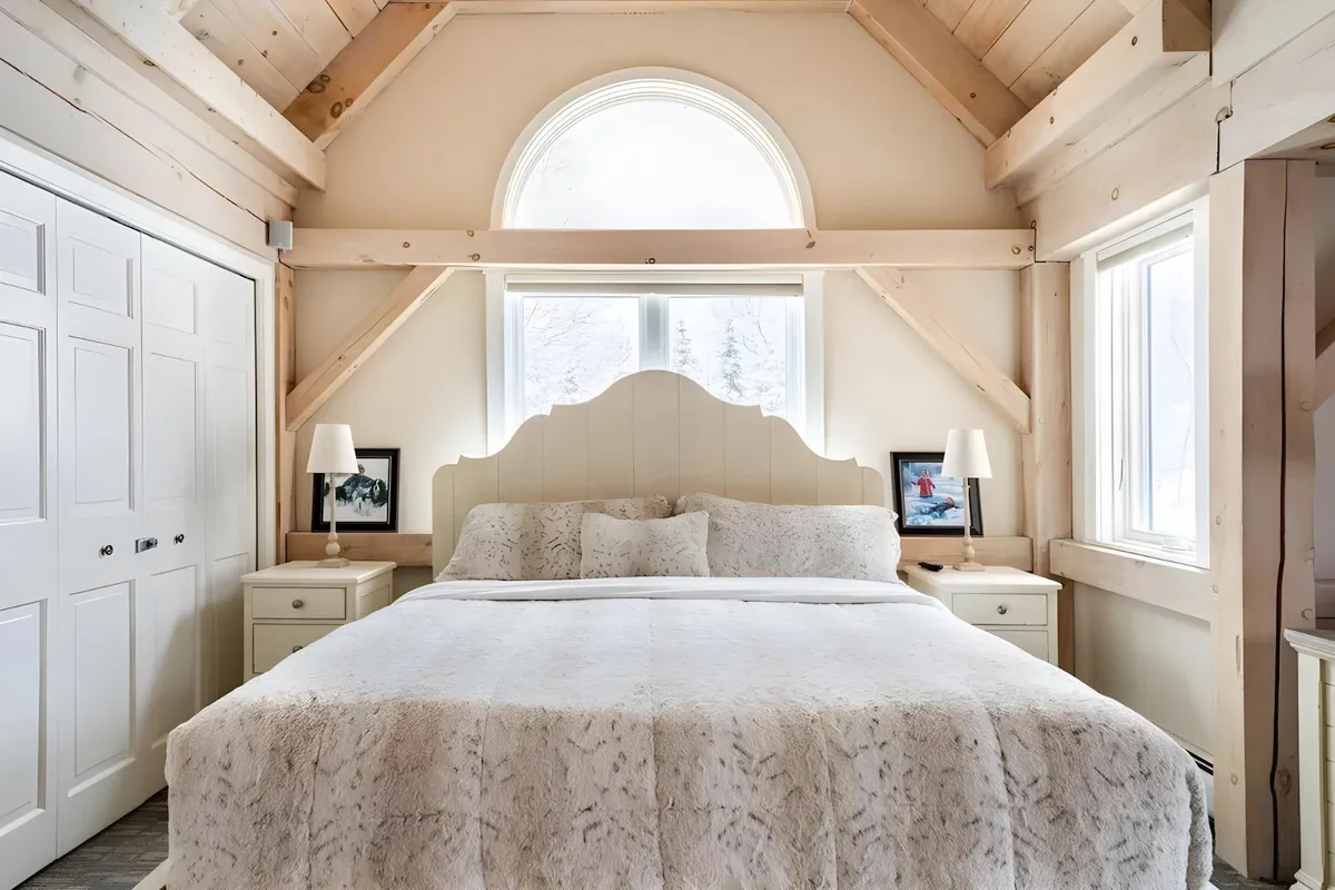 A serene bedroom centered on a large bed with plush, inviting bedding, flanked by nightstands and softly lit lamps. An arched window lets in natural light and offers views of the surrounding landscape, enhancing the room's restful atmosphere.