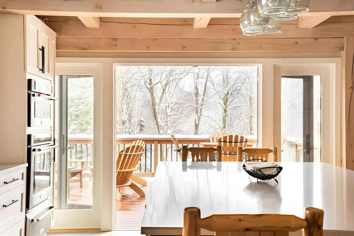 A large dining table surrounded by wooden chairs sits beside panoramic windows, offering views of a snowy landscape outside. The warm interior lighting and inviting table setting create a welcoming space for communal meals and gatherings.