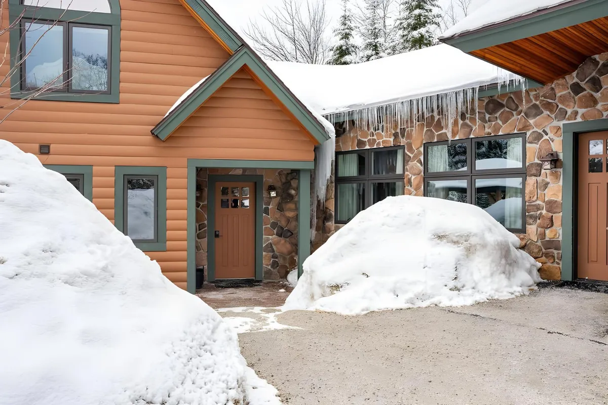 A grand stone façade stands out amidst a snow-covered driveway, icicles hanging from the roof, creating a welcoming entrance scene. The image signals arrival and accessibility no matter the season, with a sense of anticipation for the ski and après-ski experiences awaiting inside.
