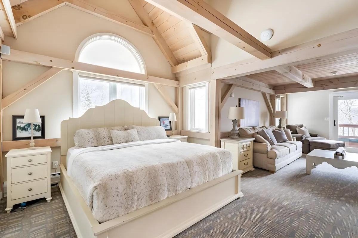 A serene bedroom and lounge area with a large, inviting bed and a comfortable sofa positioned beneath wooden beams. Expansive windows provide views of the outdoors and fill the space with natural light, creating a tranquil atmosphere ideal for unwinding with both two- and four-legged friends.
