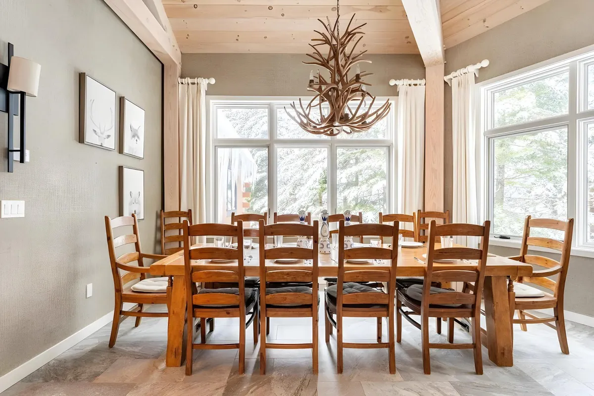 A spacious dining area with a large wooden table beneath a beautiful chandelier, set in front of expansive windows that frame breathtaking mountain vistas. The scene is set for family gatherings or festive dinners, with an atmosphere that's both elegant and welcoming.
