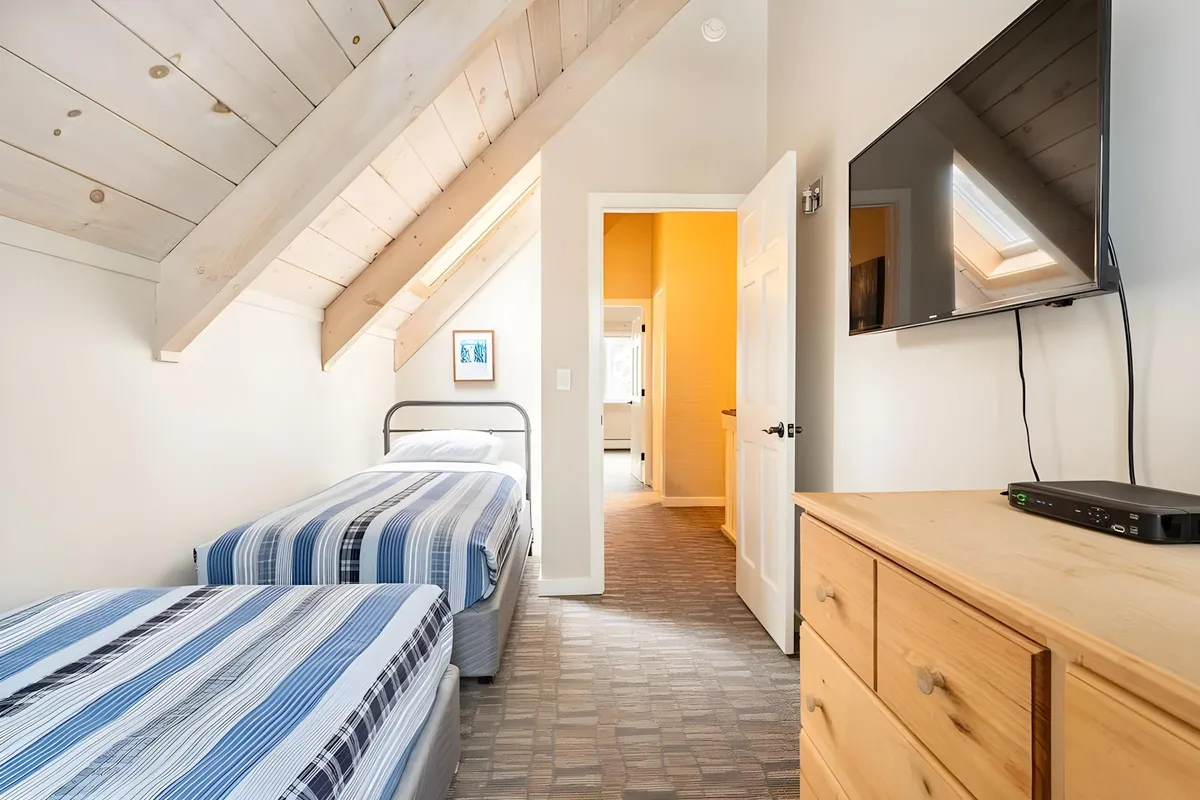 A cozy bedroom with two twin beds dressed in striped linens, a wooden dresser, a large TV mounted on the wall, and framed artwork. Natural light floods the space, creating an inviting niche ideal for flexible sleeping arrangements.