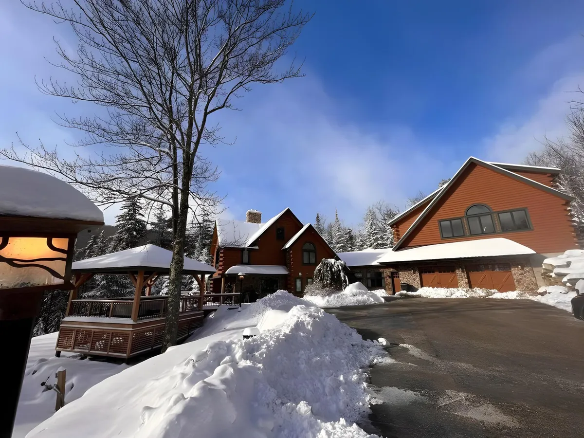 The image showcases the elegant main house nestled amidst snow-draped trees, with a pristine pathway leading up to the door, warmly lit by a classic lamp post. The scene exudes winter charm and tranquility, inviting viewers to imagine cozy family moments and outdoor fun against a breathtaking mountain backdrop.