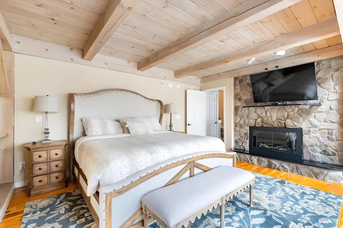 A luxurious bedroom featuring a large bed with plush bedding, a stone fireplace, TV, decorative rug, and exposed wooden beams. The room offers a cozy seating area, perfect for reading or relaxing by the fire, with ambient lighting accentuating the intimate retreat atmosphere.