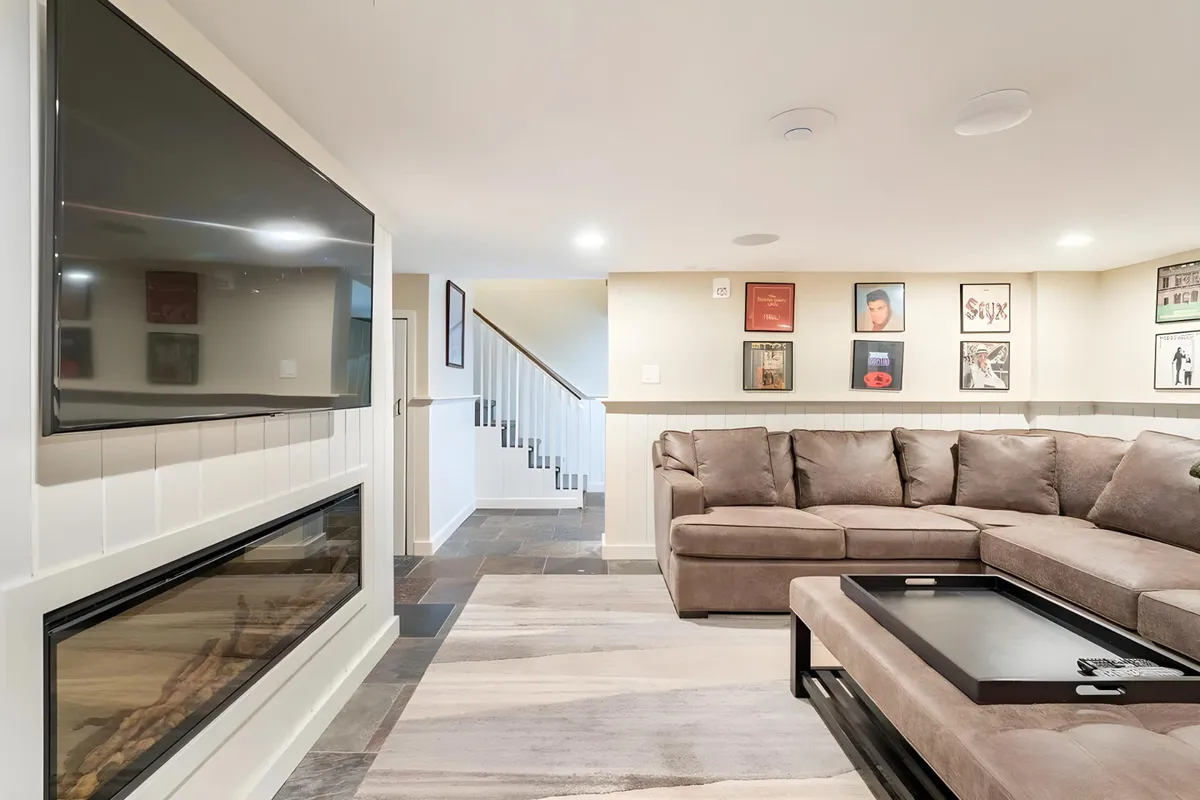 A spacious executive video room featuring a large flat-screen TV above a cozy fireplace, a comfortable sectional sofa, and tasteful artwork on the wall. The setting is perfect for group movie nights or watching sporting events, with a warm, social atmosphere.