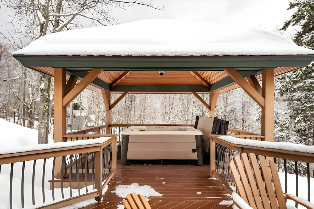 An inviting outdoor deck with a private hot tub set beneath a wooden gazebo, surrounded by snow-covered scenery. Comfortable seating areas are available for guests to relax, entertain, or enjoy the winter views while soaking in the hot tub.