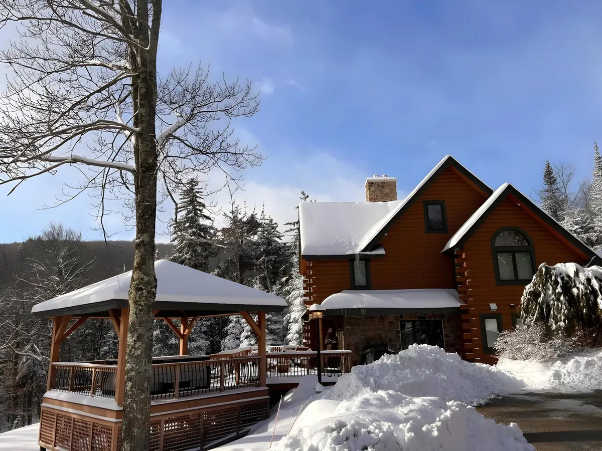 A quintessential log cabin-style house is nestled among snow-covered trees, with a wooden gazebo visible to the side. The entire landscape is blanketed in fresh white snow, evoking a classic winter retreat. The inviting exterior and natural surroundings highlight the immediate connection to outdoor activities and ski adventure.