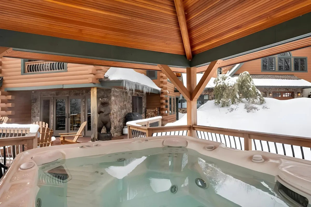 An exterior scene highlighting a private hot tub surrounded by wooden beams and a snow-covered landscape. A stone wall with a bear sculpture adds a touch of rustic charm. The image conveys relaxation and exclusivity, underscoring the private amenities mentioned in the section, such as the hot tub available only to main house guests.