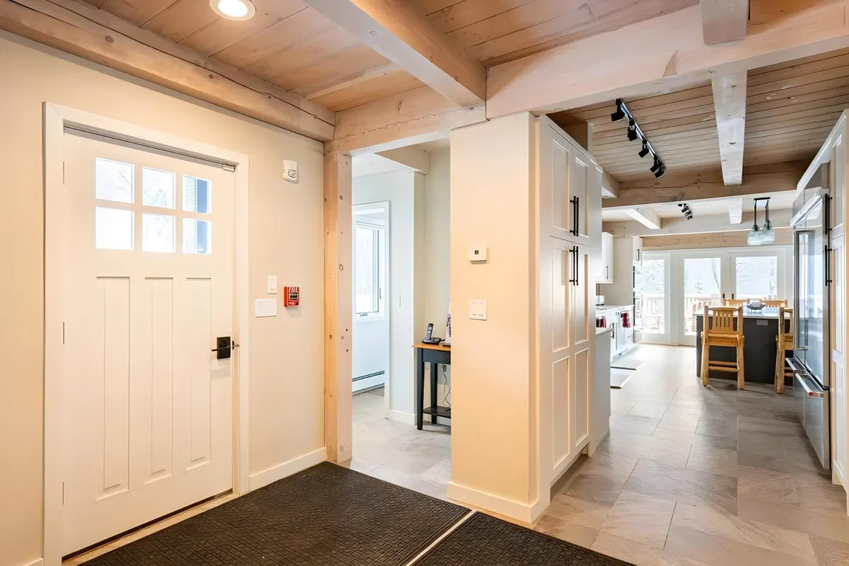 This image features a welcoming entryway with a prominent front door, wooden beams, kitchen area, and dining table visible. The space conveys a sense of arrival and comfort, suggesting easy transitions between indoors and outdoors—ideal for ski-in/ski-out experiences. The visible front door and open layout make it suitable for highlighting smart lock entry and seamless check-in.