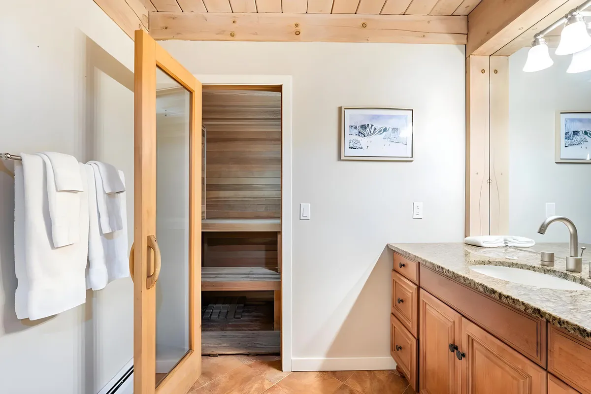 A modern bathroom with a glass shower door, a wooden sauna door, granite countertop, and towels on a rack. The space feels serene, combining spa-like amenities with natural textures for a rejuvenating experience.